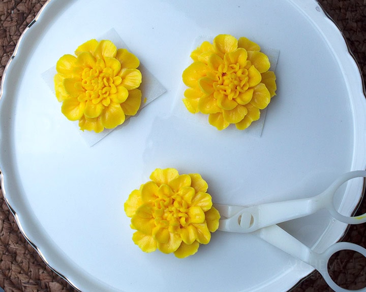 yellow buttercream marigolds arranged on a white cake stand with a pair of plastic flower scissors.