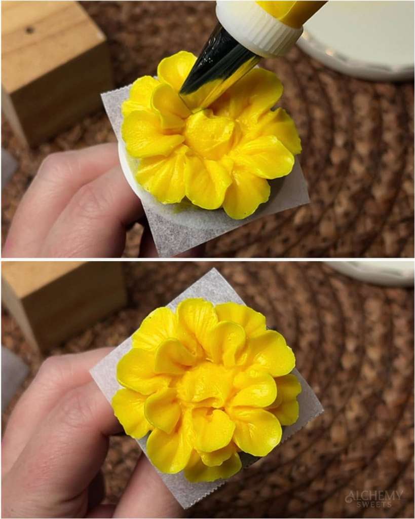 A rounder, fuller yellow buttercream marigold held in fingers after piping a third layer of petals toward the domed center.